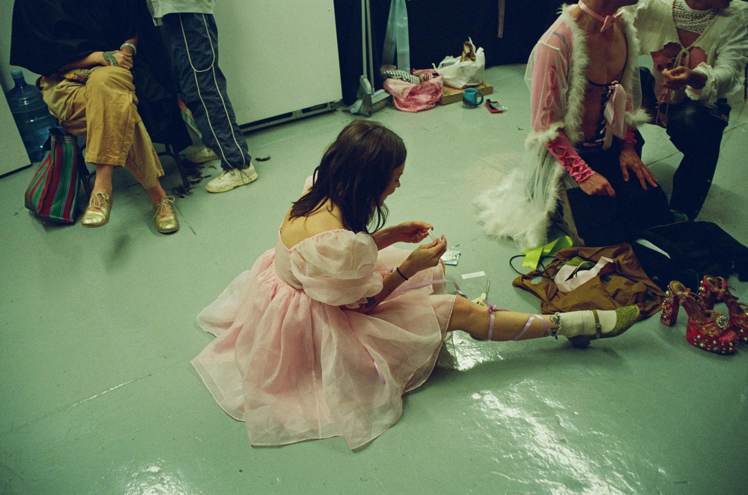 A performer in a pink tulle dress lacing her shoes on the dressing room floor